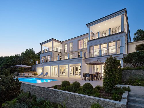 Luxuriöses Haus in Holzbauweise Eine moderne Villa mit mehreren Etagen, großen Fenstern und einem Swimmingpool im Vordergrund. Auf der Terrasse stehen Tische und Stühle, umgeben von Pflanzen und einem Garten. Der Himmel ist blau, es ist Abenddämmerung.