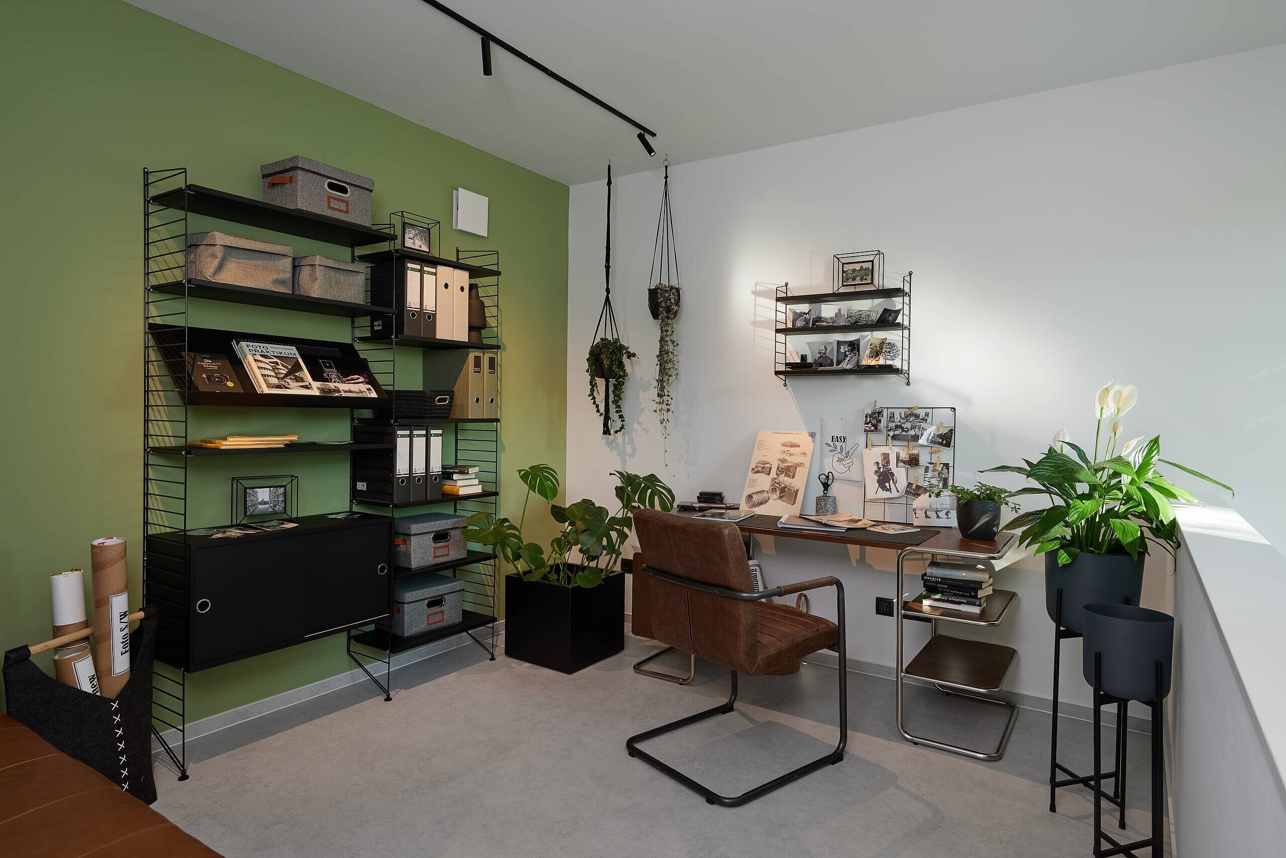 Homeoffice auf der Empore im Obergeschoss Ein helles Arbeitszimmer mit einer grünen Wand. Links steht ein offenes Regal mit Archivboxen und Büchern. Rechts befindet sich ein Schreibtisch mit einem braunen Stuhl, umgeben von Pflanzen und einer Wanddekoration aus Fotos. Die Atmosphäre wirkt modern und einladend.