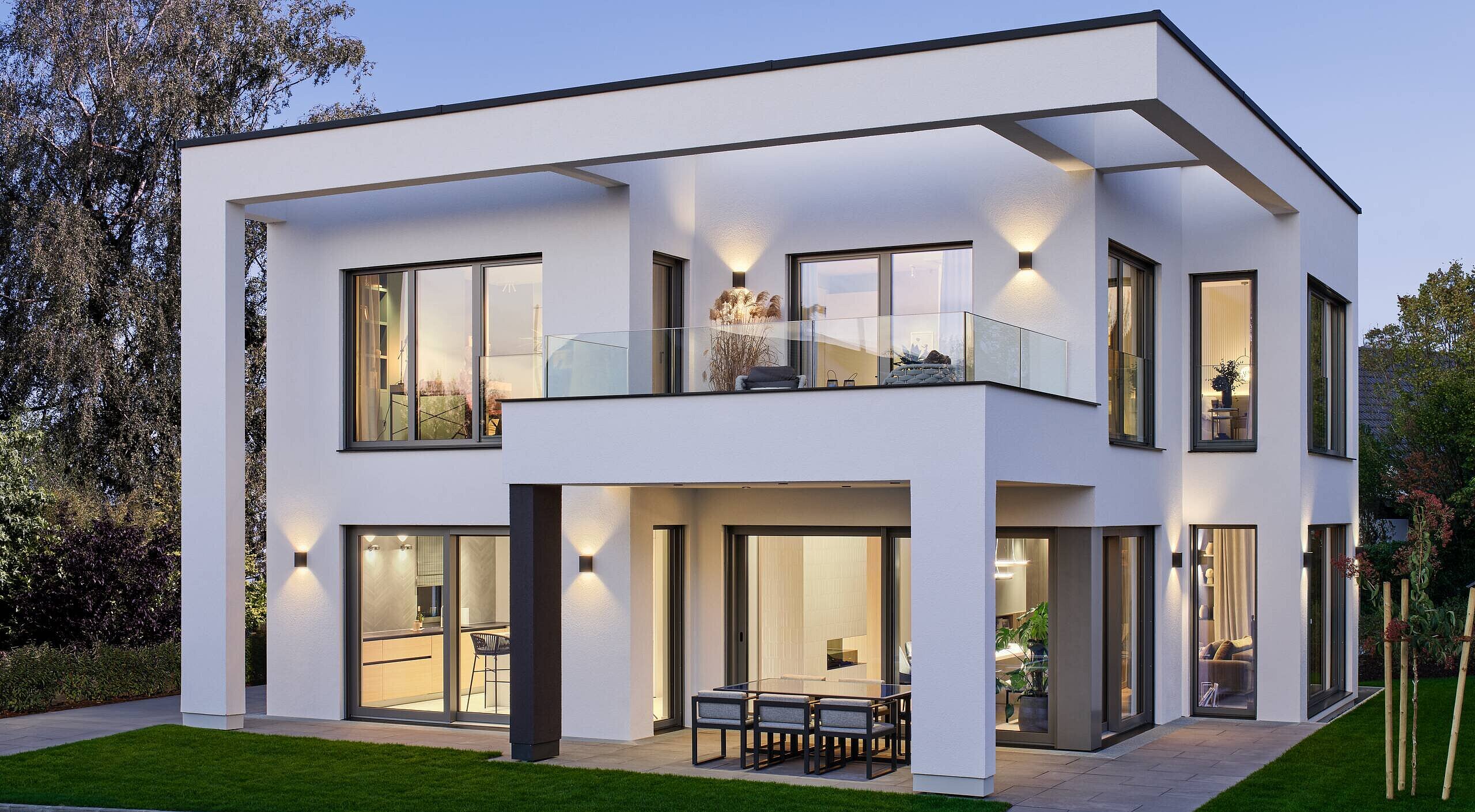 Modernes Ausstellungshaus der Luxusklasse Ein modernes, zweigeschossiges Wohnhaus mit flachem Dach. Es hat große Fenster, die Licht hereinlassen, und einen Balkon. Die Fassade ist hell und schlicht, umgeben von einem gepflegten Garten mit Rasen und wenigen Pflanzen. Der Himmel ist leicht blau, was eine angenehme Abendstimmung vermittelt.